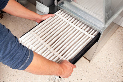 Male hands are installing a furnace air filter. This filter is not brand new but it is relatively clean, it was removed for inspection. This would be a typical filter style found in a high end newer house. The floor is epoxy painted concrete with traction chips and a floor drain.